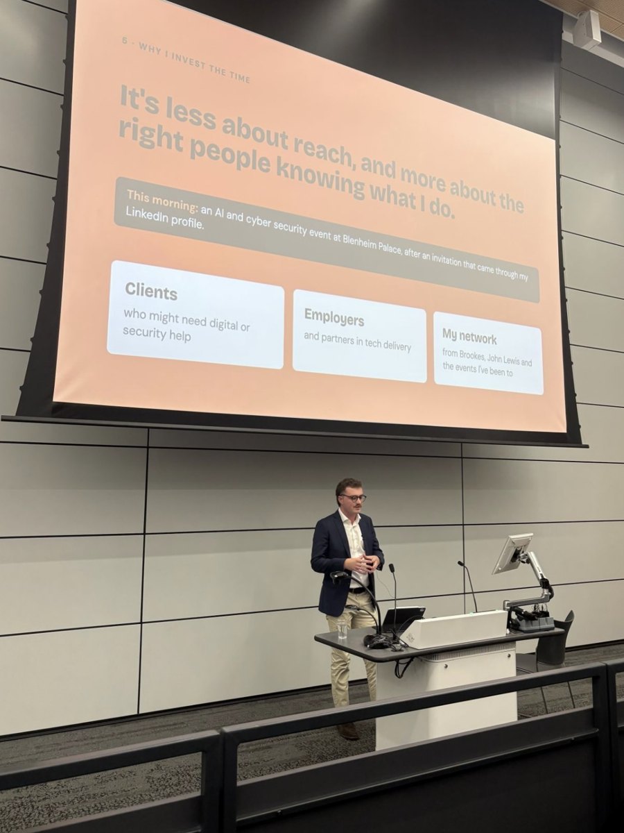 Hugo King speaking at a lectern in a lecture theatre, beneath a slide about the right people knowing what he does